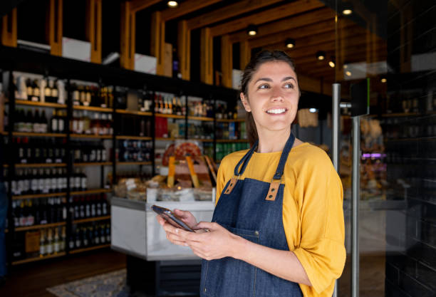 retail clerk working at a food shop and using her cell phone while waiting for customers - straatverkoper stockfoto's en -beelden