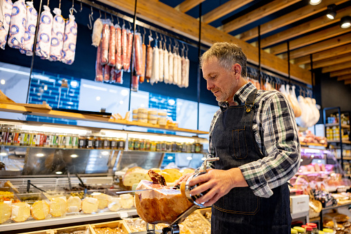 Latin American man working at a charcuterie selling hams and cutting a slice of prosciutto