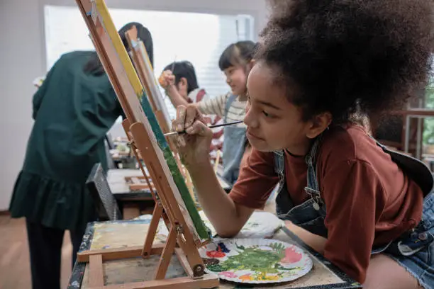 African American girl concentrates on acrylic color picture painting on canvas with students group in art classroom, creative learning with talents and skills in elementary school studio education.