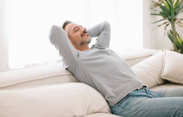 istock Relaxed man enjoying rest lying on comfortable couch with the hands on the head in living room at home. Calm middle-age man breathing fresh air enjoying no stress free peaceful weekend in house. 1438530975