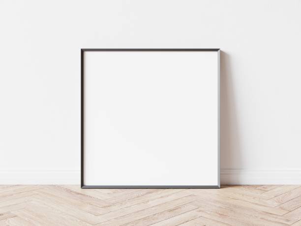 istock Empty square picture frame with thin dark border laying on wooden parquet floor, white wall in background. 3D illustration. 1438541829