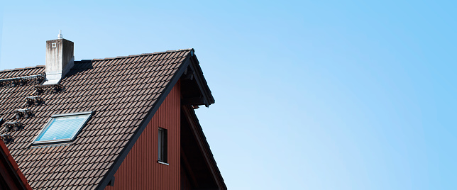 Smokestack, roof window and wooden facade of a country cottage against the sky. Plastered chimney pipe on tile roof of a private house. The exterior of the Individual heating system or ventilation on a horizontal banner