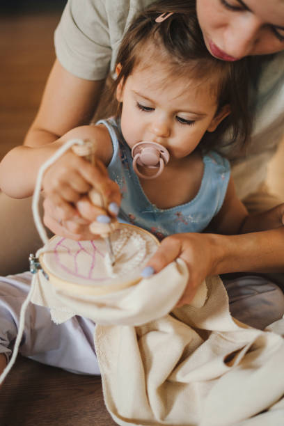 Proud mother sitting on the floor with her baby daughter, embroidering on a loop together. She's hugging and encouraging her. Colorful pattern. Home decoration. stock photo