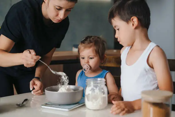 young mother with her two kids weighing out flour into bowl in kitchen environment, baking cakes for the party. Weighing indigrients on electronic kitchen scales