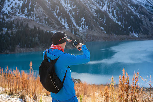 Warmly dressed man taking a picture with his phone of the stuningly beautiful lake surrounded by snowy mountians. Dry grass in foreground.