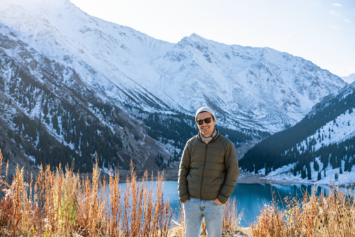 Portrait of a smiling man in sunglasses and warm clothes over lake surrounded by snowy mountians. Dry grass in foreground.