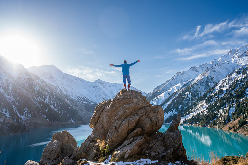 Longshot of a man standing on a huge boulder on a vantage point over stunning turquoise lake surrounded by snowy mountains on a sunny day. His hands are outstretched like wings.