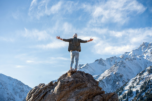 Hyped man standing on a rock surrounded by snowy mountains and blue sky on a sunny day.
