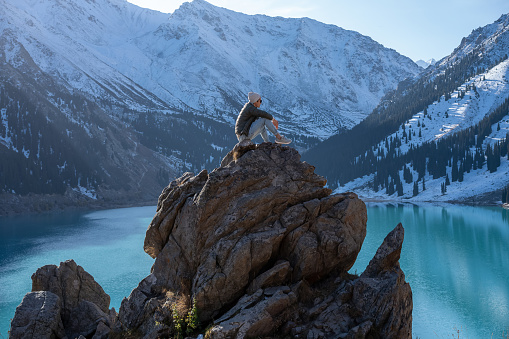 Side view of a man sitting on a huge boulder on a vantage point over stunning turquoise lake surrounded by snowy mountains on a sunny day.