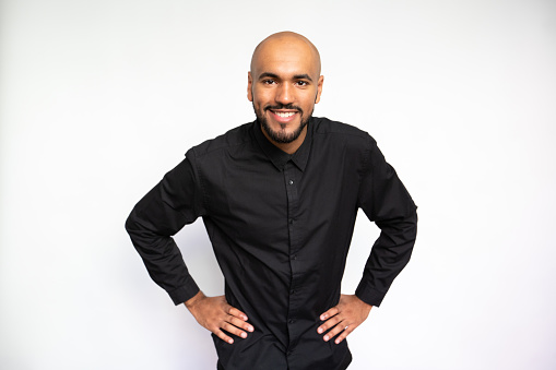 Portrait of excited young man with hands on waist. Happy multiethnic male model with bald head and beard in black shirt looking at camera, bending and smiling. Self-assurance, success concept.