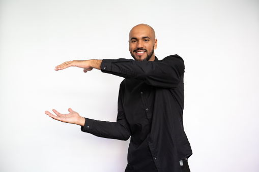 Portrait of happy young man holding invisible object. Excited Latin American male model with bald head in black shirt looking at camera, smiling while showing ads. Advertisement, size concept