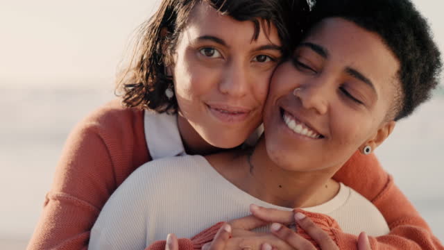 Couple, lgbt and lesbian women at the beach hugging, embrace and holding each other. Love, romance and   portrait of lesbian couple enjoying sunset, holiday and summer by the ocean with smile on face