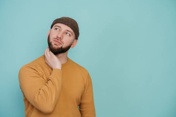istock Thoughtful young beardy man in hat and sweater looking aside in dreaming expression over turquoise background. Hesitated Caucasian guy thinking, makes a choice. Mockup. Handsome guy makes decision. 1438569652