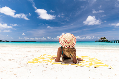 Back view of a woman with sun hat relaxing on a beach towel in summer day and looking at view. Copy space.