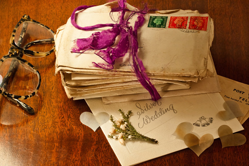 A bundle of old love letters with King George VI postage stamps from the late 1930's and tied with a fraying ribbon, accompanied by Wedding and Wedding Anniversary memorabilia, including a Post Office telegram a silver wedding anniversary card, and a 'good luck token' of a sprig of white heather: all evoking fond memories of an enduring love. Memories brought back to life by the reader's spectacles.