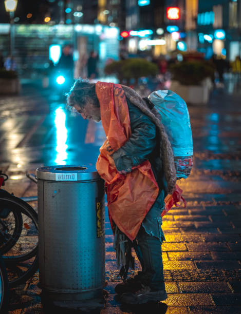 Munich, Germany – February 01, 2020: Homeless man looking for drinks and food in garbage bin. Cold rainy night in Munich with wet streets reflecting the lights of the city