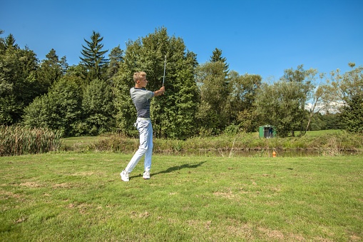 A great shot of a young man playing golf in a field surrounded by trees on a sunny day