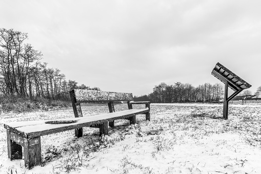 A grayscale shot of benches on a field covered in snow under a cloudy sky