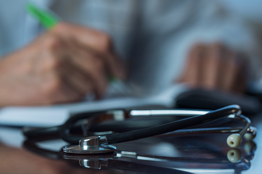 Medical stethoscope lying on desk against blurred doctor hands with pen and writing recipe