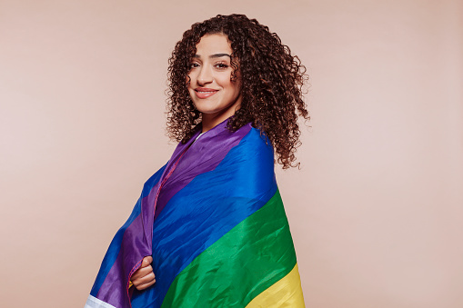 Portrait of happy arabic woman with curly hair in multi color rainbow flag at studio isolated over beige background. Homosexual, lesbian, human rights, lgbt pride concept.