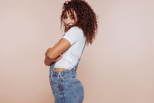 Side view studio portrait of shiny adorable girl with curly hair, dressed denim overalls and white t-shirt, with crossed arms, empty space, isolated over beige color background.