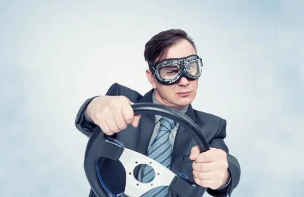 Photo of Confident businessman in a dark suit and tie, in stylish goggles, turns a car steering wheel, on a light blue smoky background