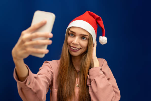 A girl in a Santa Claus hat on an isolated blue background is photographed on her phone stock photo