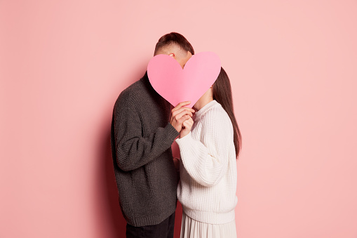 Portrait of lovely young couple, man and woman kissing behind paper heart, posing isolated over pink background. Concept of love, relationship, Valentine's Day, emotions, lifestyle, holiday. Ad