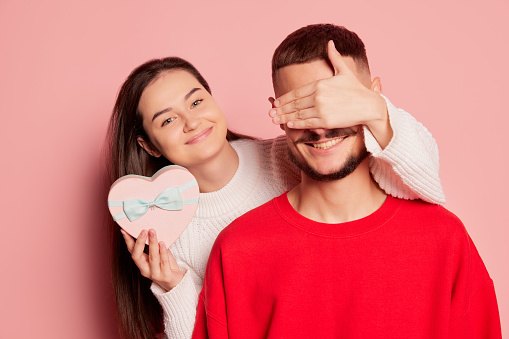 Portrait of young couple, beautiful woman covering man's eyes and giving present isolated over pink background. Holiday surprise. Concept of love, relationship, Valentine's Day, emotions, lifestyle