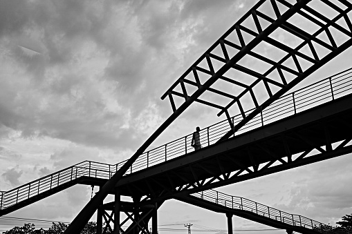 A greyscale low angle shot of a male walking through a bridge under the cloudy sky