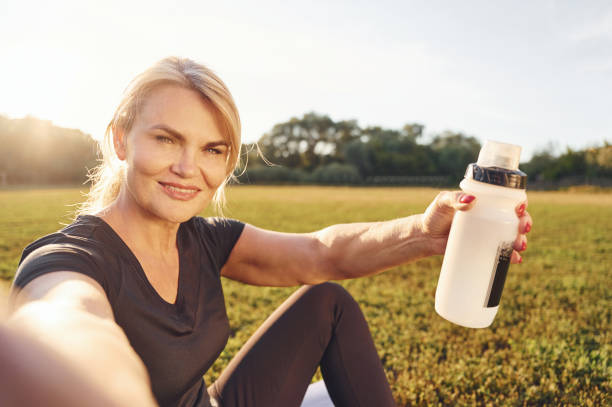 Making selfie. Holding bottle of water. Mature sportive woman in fitness clothes is outdoors at daytime stock photo
