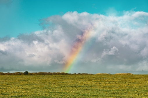 A beautiful shot of an empty grassy field with a rainbow in the distance under a blue cloudy sky in England