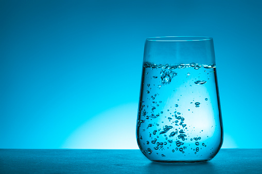 Front closeup view of a glass full of water and bubbles. The glass is at the right side of the image leaving a useful copy space at the left side on a gradient blue background.