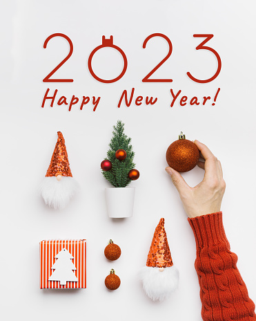 New Year holiday flat lay, shiny red Christmas ball in woman hand. Festive Christmas pattern with paper gifts, dwarfs with red shiny cap, fir tree, text 2023 Happy New Year. Winter Holiday greetings