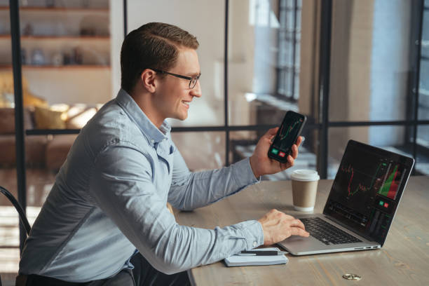 istock Smiling crypto trader with phone checking candlestick graph on laptop, buying and selling currency 1438621702