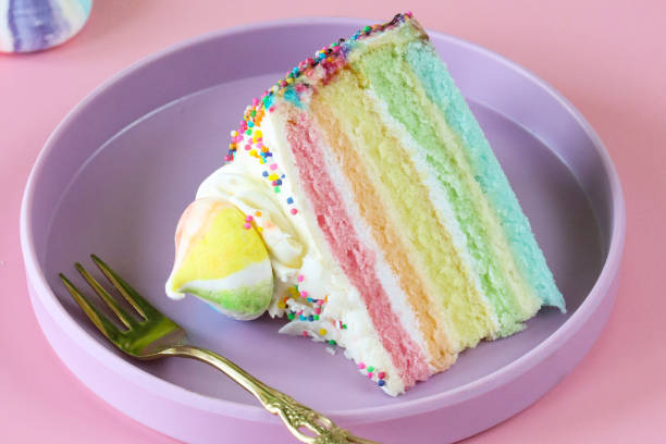 istock Close-up image of rainbow cake slice, five rainbow sponge layers separated by vanilla buttercream on purple plate with fork, piped icing rosette, rainbow meringue kiss, pink background, focus on foreground 1438625752