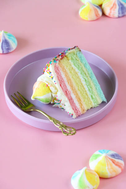 Image of rainbow cake slice, five rainbow sponge layers separated by vanilla buttercream on purple plate with fork, piped icing rosette, rainbow meringue kiss, pink background, focus on foreground