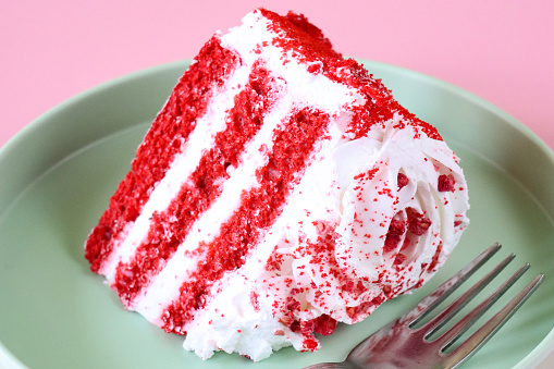 Stock photo showing close-up view of a slice of red velvet cake displayed on a plate against a pink background.