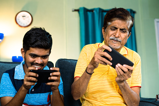 Excited father and son playing video game on mobile phone at home while sitting on sofa - concept of entertainment, technology and family