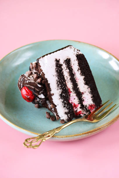 istock Image of Black Forest gateau slice, three cake layers separated by whipped cream on duck egg blue plate with fork, piped buttercream rosette, glace cherry, luxury chocolate cake, chocolate shavings, pink background, focus on foreground 1438625972