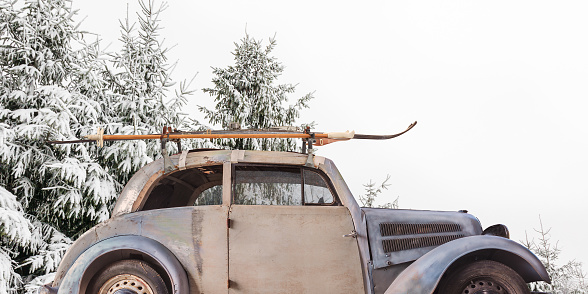 Vintage weathered car with skis attached to a roof rack in front of snow covered fir trees