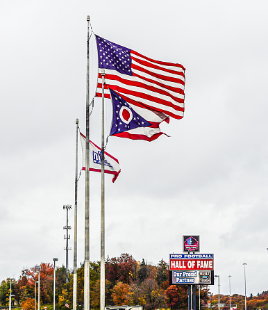 Canton, Ohio, USA---October 26, 2022: A Pro Football Hall of Fame flag under an American flag and Ohio State flag, waving from flag poles in Canton, Ohio.   Pro Football Hall of Fame signs in the background. Close-up.