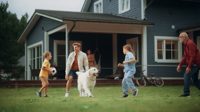 Joyful Young Kids and Adults Playing Ball with an Energetic White Golden Retriever. Cheerful People Playing Football with Pet Dog on a Lawn on Their Front Yard in Front of the House.