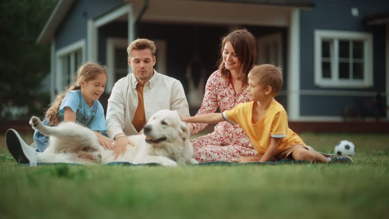 Portrait of a Cheerful Family Couple with Kids, Playing and Petting a Beautiful White Golden Retriever Dog. Happy Successful People Sitting on a Lawn in Their Front Yard in Front of the House.