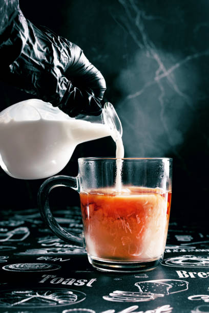 Tea with milk, milk in motion from a gravy boat pours into a cup, steam from a hot drink, a dark cold background, a hand in a black glove stock photo