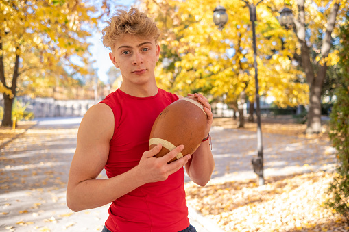 Active young boy with blond hair, blue eyes, a red tank top, and an American football ball in a public park.