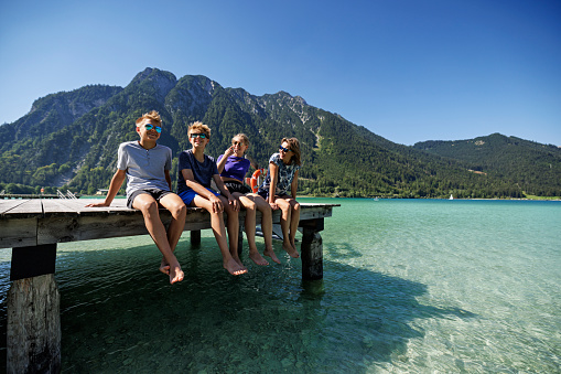 Mother and teenage kids enjoying summer vacations. They are sitting on pier on a lake Achensee in Austrian mountains. \nSunny summer day.\nCanon R5