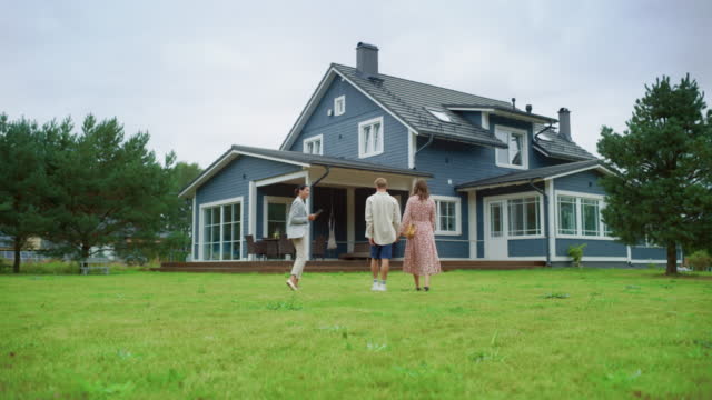 Establishing Shot: Professional Real Estate Agent Showing a Hot Property Location to a Young Successful Couple. Female Real Estate Agent with Tablet Walking with Family to the House. New Homeowners. Slow Motion