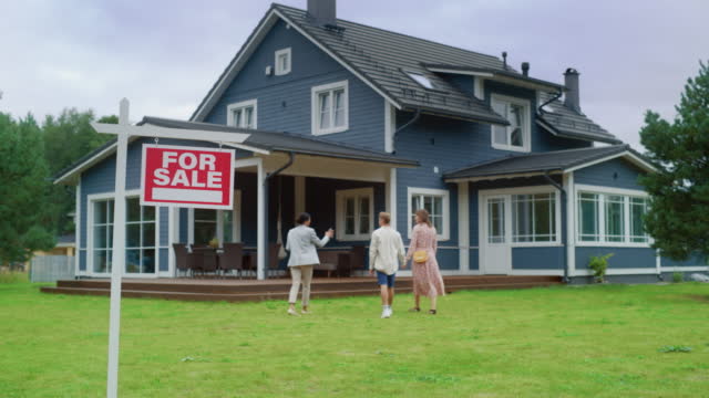 Real Estate Agent Meeting a Beautiful Successful Couple in Front of a Modern Big House That is For Sale. Real Estate Agent Selling or Renting Out a New Home to a Young Family. For Sale Sign on Front Lawn.