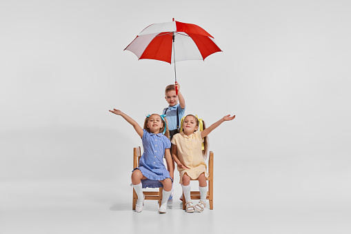 Portrait of three children, boy and girls playing together, sitting on chair with umbrella isolated on grey background. Having fun. Concept of childhood, creativity, retro vintage fashion, friendship